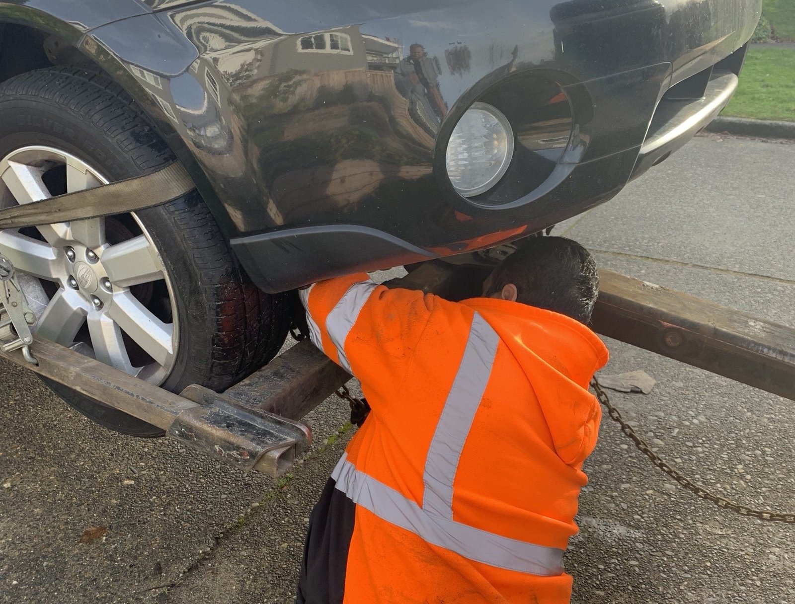 A tow driver in a high-visibility vest kneeling at the wheel of a car on the roadside, hooking it up, with traffic lanes behind.